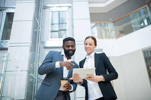 Positive Black manager with coffee cup pointing at tablet while discussing sales plan with colleague