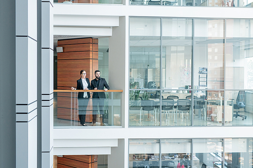 Young employees of business center standing at railing and looking around, interior of glassy office room