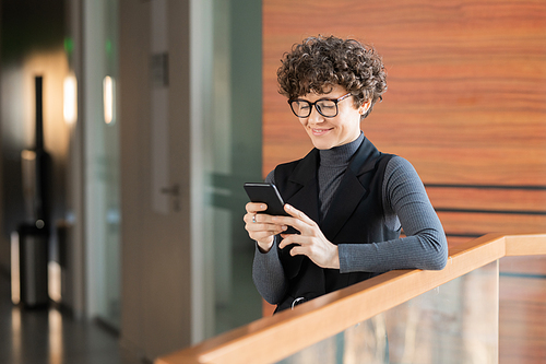 Smiling young curly-haired lady standing in corridor and checking social media statistics on phone