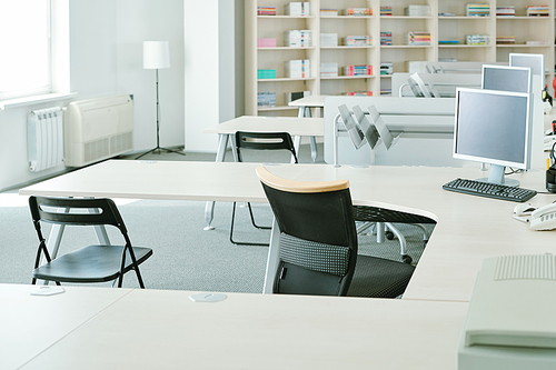 Row of desks with computer monitors and chairs near by on background of shelves with documents and supplies and lamp by window in office