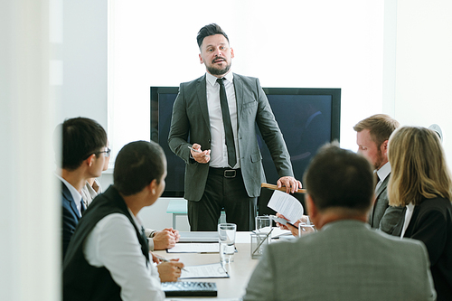 Confident middle aged businessman in formalwear standing by table in front of his colleagues and making report or speech at training