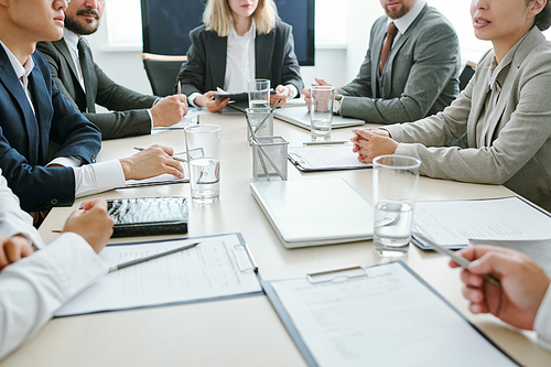 Mid section of contemporary intercultural brokers or business partners sitting around table in boardroom and discussing strategies