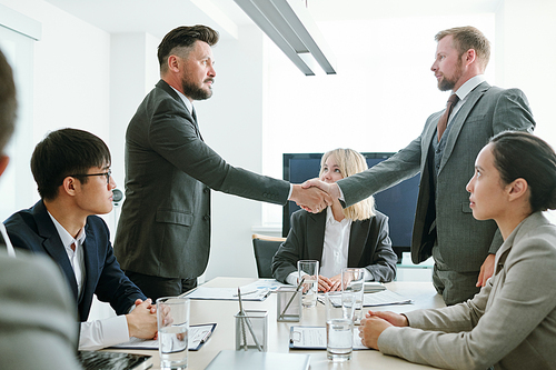 Two serious business partners in formalwear handshaking over table among their colleagues after negotiating and signing new contract