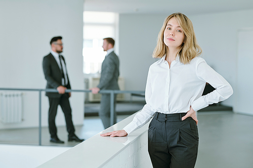 Young blond successful businesswoman in formalwear keeping hand on waist while standing in front of camera against two male colleagues