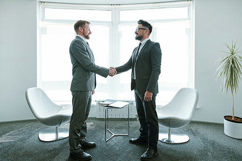 Two young successful businessmen in suits shaking hands after negotiation while standing against office window, table and armchairs