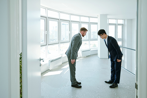 Young Chinese and Caucasian businessmen in formalwear making bow to one another during greeting while standing inside large office building