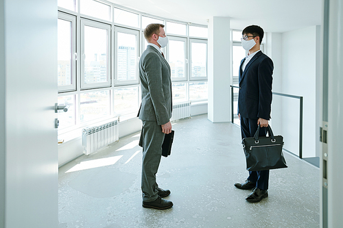 Two young contemporary businessmen in elegant suits and protective masks standing by large office window in front of one another