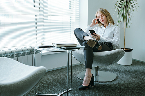 Young blond relaxed businesswoman in formalwear sitting in armchair by large office window and scrolling in smartphone at break