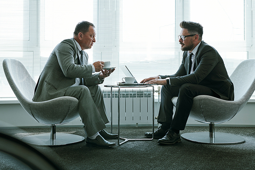 Two elegant businessmen in suits sitting in armchairs against large office window and discussing terms of contract or new strategies