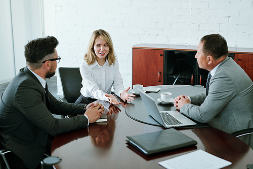 Young blond businesswoman looking at one of male colleagues while explaining her viewpoint or idea at start-up meeting in office