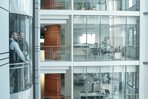 Two young businessmen moving in elevator inside large contemporary business center consisting of many floors