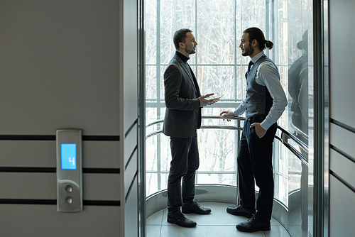 Two elegant businessmen discussing working points or terms of agreement inside elevator of large business center