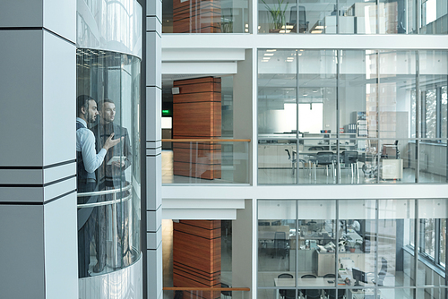 Two young intercultural managers having talk while moving upwards in elevator inside large modern business center with offices