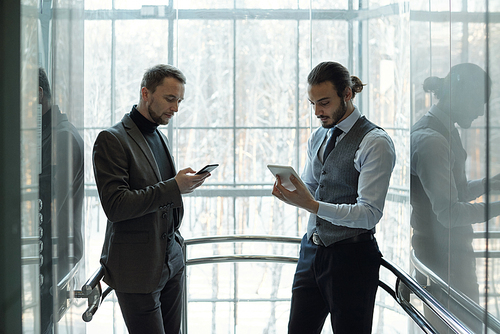 Two young well-dressed businessmen using mobile gadgets at break while standing by window of modern business center