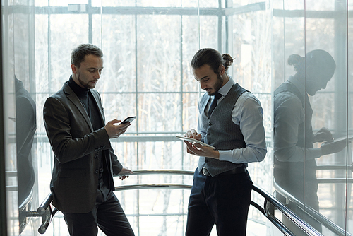 Two young serious businessmen in formalwear using mobile gadgets while moving in elevator inside contemporary business center