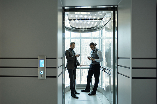 Two young well-dressed male entrepreneurs using mobile gadgets in elevator of modern business center