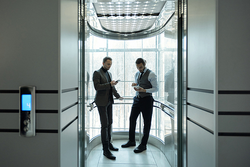 Two young elegant office managers scrolling in mobile gadgets while standing in elevator with open door inside business center