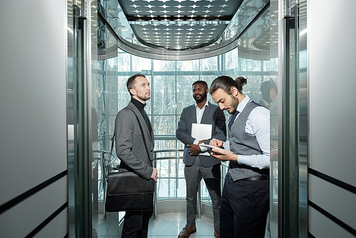 Young well-dressed male entrepreneur using tablet in elevator of modern business center among his colleagues