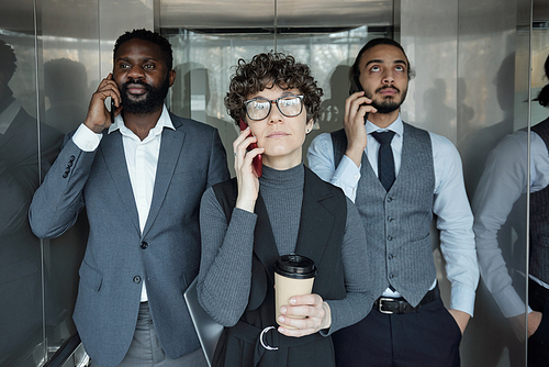 Young businesswoman with glass of coffee and two intercultural male colleagues calling by smartphones in elevator