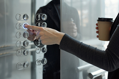 Hand of young businesswoman with drink pushing button inside elevator while going back to office on fifth floor at coffee break