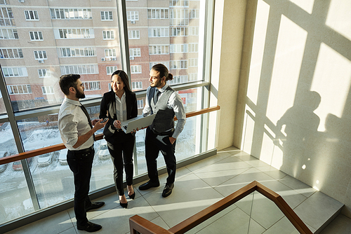 Group of three young contemporary analysts in formalwear discussing online data at meeting by window