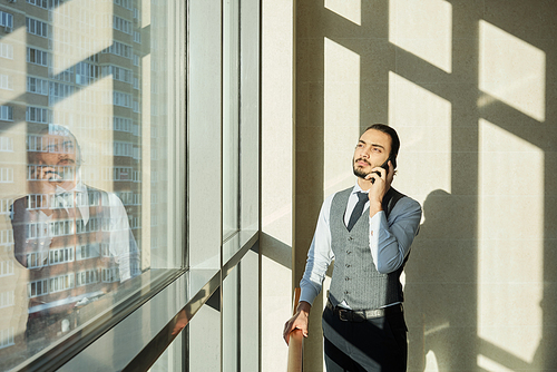 Young elegant businessman standing by window of office center on sunny day and talking to client by mobile phone