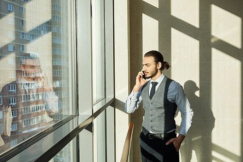 Young serious businessman looking through window on sunny day while consulting one of clients by smartphone on working day