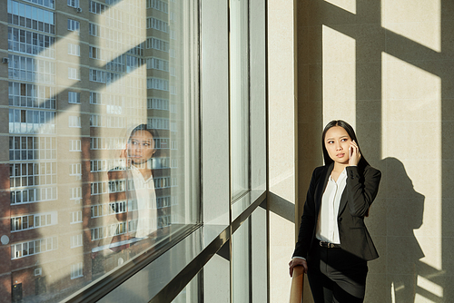 Pretty young Asian businesswoman standing by window of office center and talking to client by mobile phone