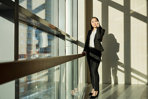 Pretty young mixed-race businesswoman in elegant suit standing by large window inside business center and talking on mobile phone