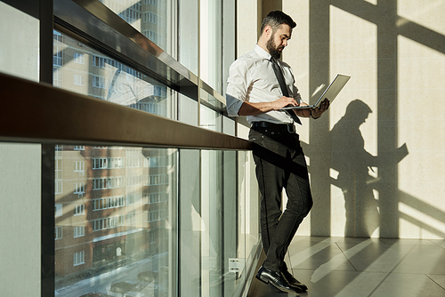 Young elegant businessman with laptop standing by window of office center while surfing in the net for online data