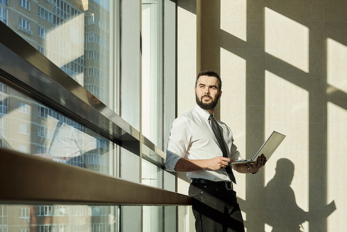 Young pensive businessman with laptop preparing presentation or working over new business project by window in office center