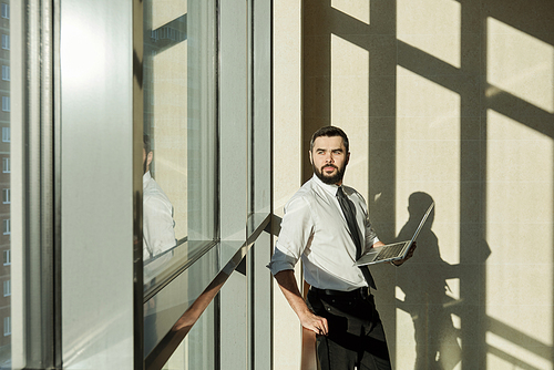 Young elegant businessman with laptop looking through window of office center while browsing in the net