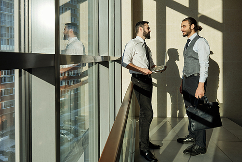 Two young successful businessmen standing by large window during discussion of working details at meeting in corridor