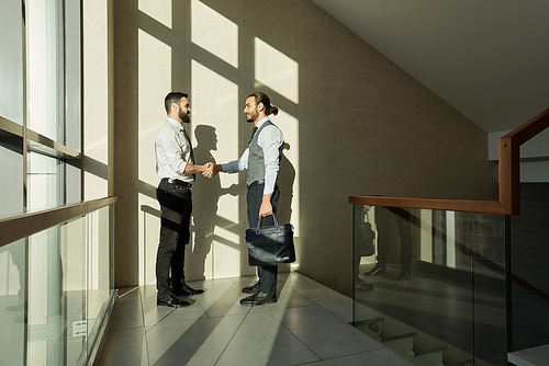 Two young elegant business partners handshaking after negotiating and signing new contract while standing against wall