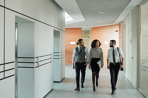 Three young well-dressed intercultural colleagues discussing working points on the move inside large business center