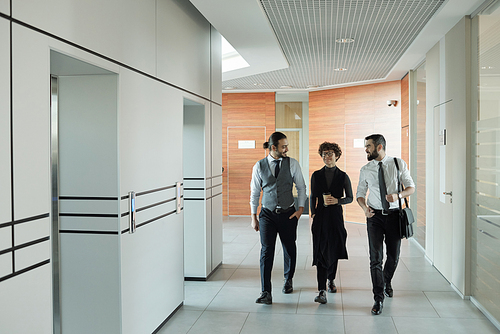 Three young cheerful colleagues in formalwear chatting on their way to office while passing by elevator after coffee break