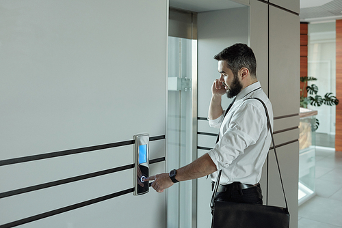 Young busy office manager with smartphone talking to client while standing by elevator and pushing call button