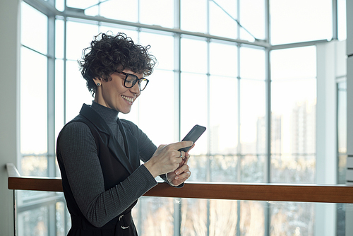 Pretty young smiling businesswoman scrolling in smartphone while standing by railings inside office center