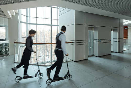 Blurred outlines of young managers moving on scooters in corridor of contemporary business center