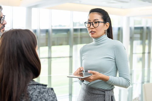 Young Asian female office manager in casualwear and eyeglasses holding digital tablet in front of herself while talking to colleague at meeting