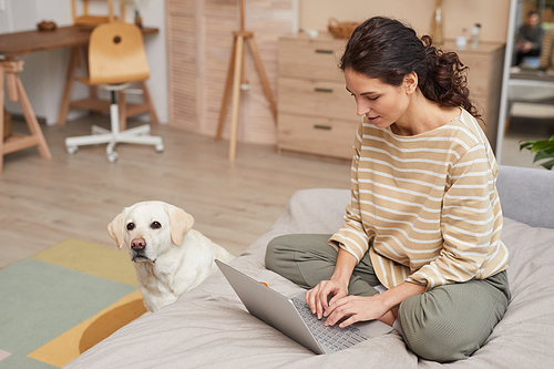 Full length portrait of smiling young woman using laptop on bed with pet dog in cozy home interior, copy space
