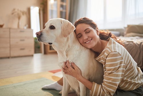 Warm toned portrait of smiling young woman embracing dog while sitting on floor and enjoying time with loving pet, copy space