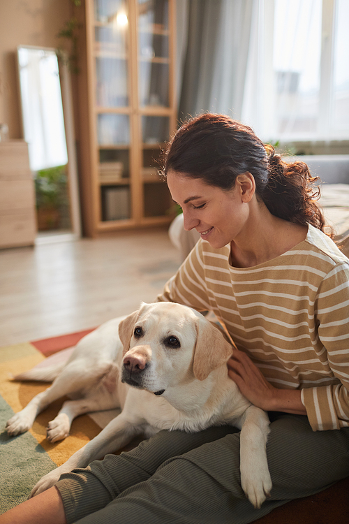 Vertical warm toned portrait of smiling young woman hugging dog while sitting on floor in cozy home interior