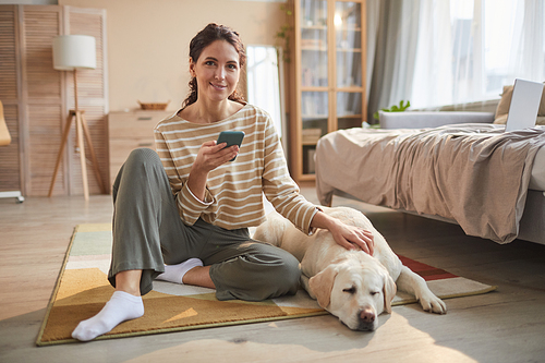 Full length portrait of smiling young woman sitting on floor holding smartphone and petting dog in cozy home interior, copy space