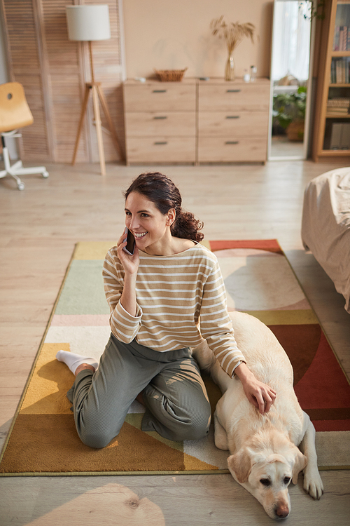Vertical high angle portrait of smiling young woman speaking by smartphone while sitting on floor with white labrador dog in cozy home interior