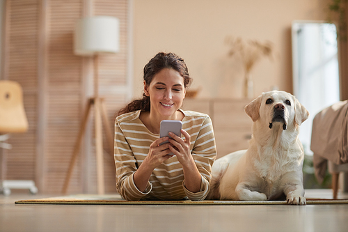 Front view portrait of smiling young woman lying on floor with dog and using smartphone in cozy home interior, copy space