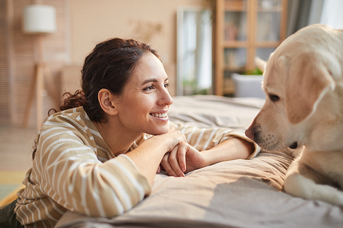 Side view portrait of smiling young woman looking at pet dog with love and care in cozy home interior, copy space