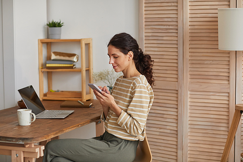 Warm toned portrait of modern young woman using smartphone at home office while enjoying remote work in cozy interior, copy space