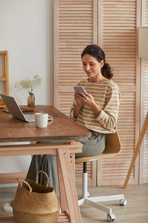 Vertical warm toned portrait of modern young woman using smartphone at home office while enjoying remote work in cozy wooden interior, copy space