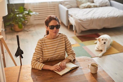 Warm toned high angle portrait of modern blind woman reading braille book while sitting at table in cozy home interior with guide dog in background, copy space